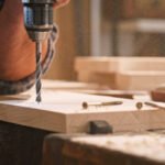 Close-up of male carpenter drilling wooden plank with drill in workshop.